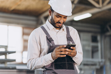 Happy bearded Indian male factory worker in special uniform and white hard hat holding the phone...