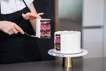 Housewife in black apron taking piece of delicious homemade cake with spatula standing at kitchen table confectioner demonstrating result of work custom pastry preparation at home