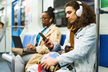 Serious teenage guy in headphones texting in mobile phone while sitting in subway train against...