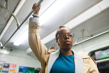 Young serious African American woman in earphones holding by handrail and looking forwards through...