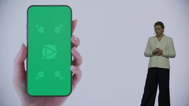 A woman presenter is standing on a well-lit stage in front of a large LED screen showing a hand holding a generic smartphone with green screen. Technology, application or product launch presentation 