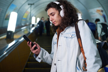 Teenage guy in casualwear and headphones using smartphone while standing on escalator, watching...