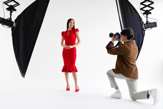 Fashion photoshoot. Professional photographer taking photos of female model in elegant red dress, posing in photo studio