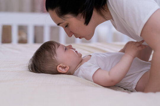 Loving mom leaning to kiss adorable little son lying on soft bed in nursery at home happy mother enjoying spending morning with chubby baby boy in white bodysuit