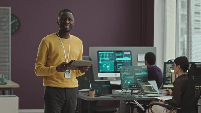 Medium portrait of young African American software engineer with digital tablet in hands smiling at camera standing in modern office with colleagues monitoring data on computers in background