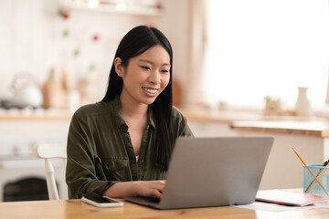 Smiling young asian woman sitting at kitchen, using laptop