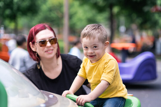 Cute little boy with Down syndrome sitting on playground equipment near mother in sunglasses looking in camera and smiling child and parent having fun on city playground