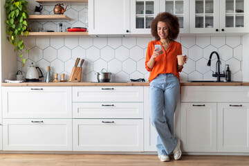 Happy Young Black Woman Using Smartphone And Drinking Morning Coffee In Kitchen