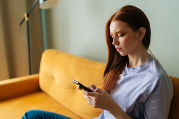 Sad red-haired young woman looking at smartphone screen expression face thinking about problem...