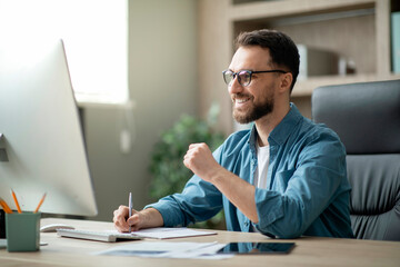 Smiling male entrepreneur using computer and taking notes at workplace in office