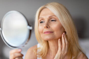 Attractive aged european woman looking in magnifying mirror and touching her face smooth skin,...