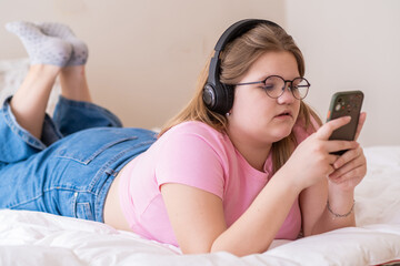 Concentrated overweight teenage girl in headphones lies on bed watching TikTok videos schoolgirl in...