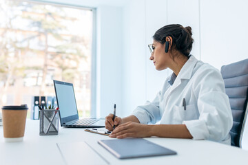 Beautiful female doctor doing a video call while talking with earphone in the medical consultation.