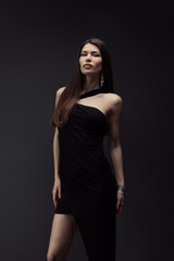 Sensual young female in stylish black dress standing in studio and looking at camera on black background during photo shoot against dark backdrop