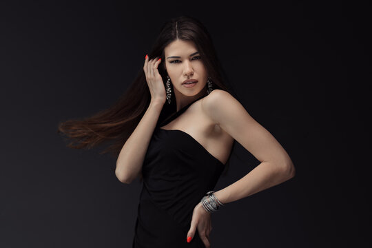 Young attractive female in black dress touching hair and posing for photo session against dark background looking at camera with toothy open mouth