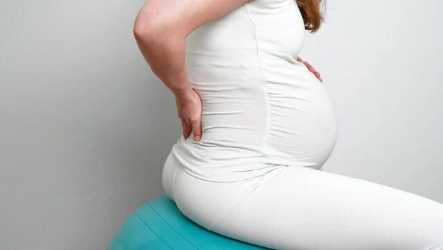 Midsection of heavily pregnant baby bump held by hands of woman bouncing on exercise ball. Side view. White background. Bright shot.