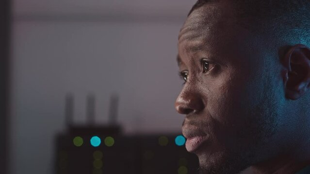 Medium closeup of focused African American male software engineer looking at computer monitor while working in dark server room