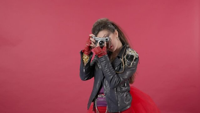 A woman sets up an old camera, points the lens and takes pictures. Woman freak, with bright makeup and bouffant in the studio on a red background. Advertising. HDR BT2020 HLG Material.