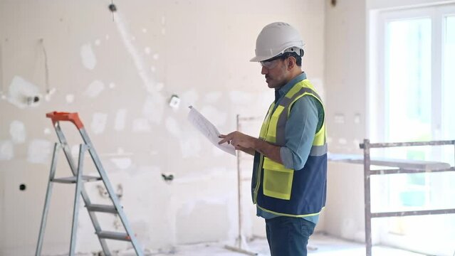 Focused architect studies apartment project drawing standing in unfinished room professional worker in protective hardhat controls renovation and construction process