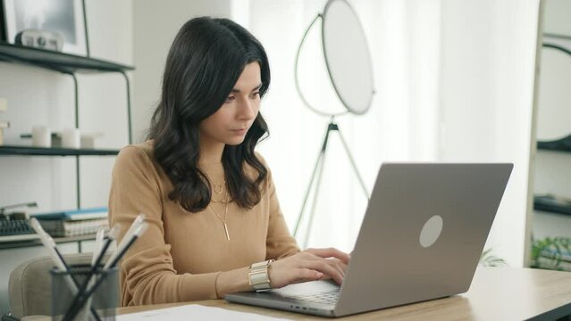Focused latin female professional writing important email in home office. Modern living room remote working. Side view dolly shot of confident hispanic brunette woman open and typing on modern laptop