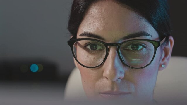 Closeup of concentrated young female programmer working on computer in dark office at night with green program code encryption reflecting in her eyeglasses