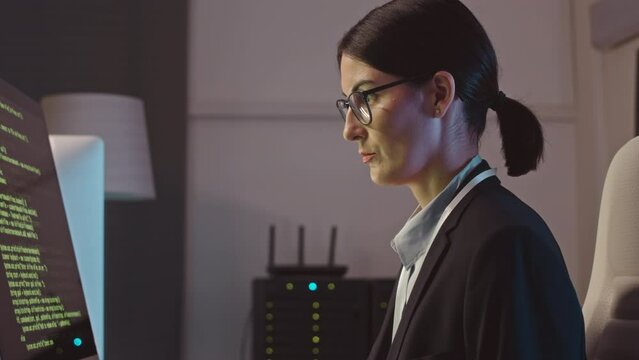 Waist up of serious Caucasian female hacker writing sophisticated program code on desktop computer while working in dark cybersecurity agency