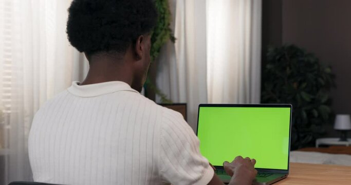 A man sits in a room at a desk and shops online. He is looking for online clothing stores. The man taps his fingers on the keyboard displayed green screen.