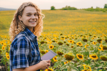 Positive female in checkered shirt smiling and looking at camera while standing in meadow with yellow sunflowers in countryside