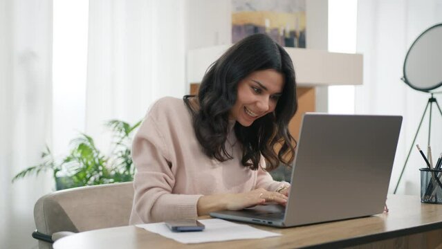 Happy girl celebrates online success, working remotely at desktop at home. Euphoric latin lady gets new distance job opportunity reads good news 4K. Excited young hispanic woman winner looks at laptop