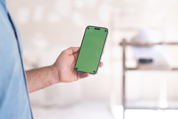 Man holding phone with green chromakey screen in hand against construction equipment in unfinished room owner ordering renovation services using mobile app