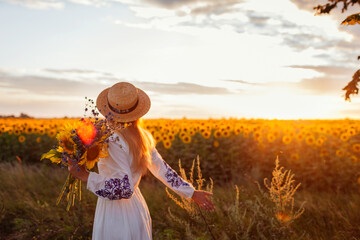 Back view of woman walking by blooming sunflower field at sunset with bouquet of flowers. Peace and...