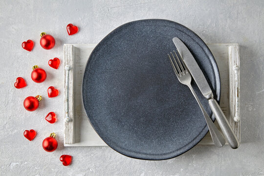 An empty gray ceramic plate on a white tray with cutlery surrounded by red christmas balls and hearts on a light concrete table