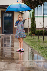 Naklejka premium Child with umbrella walking under the rain in the city, happy girl with rainbow colorful umbrella near the house, warm wet summer weather with puddles 