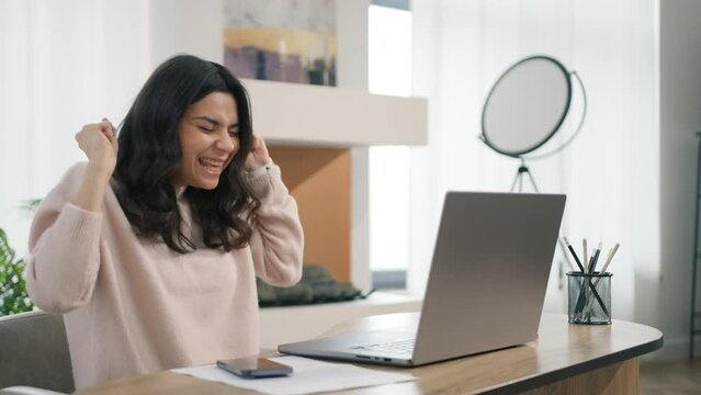 Excited young hispanic woman winner looks at laptop. Happy girl celebrates online success, working remotely at desktop at home. Euphoric latin lady gets new distance job opportunity reads good news 4K