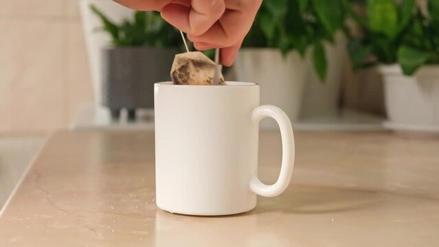 A man mixes sugar with a teaspoon in a white teacup with a tea bag and hot water to make himself tea in the kitchen. Break.