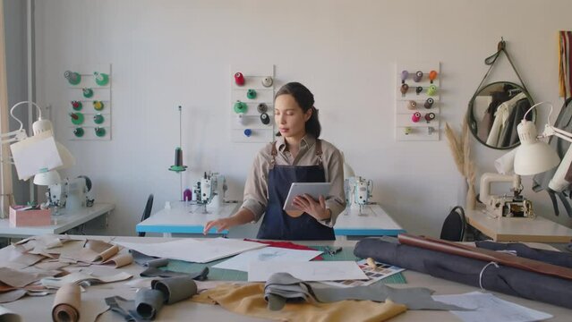 Full shot of young female biracial tailor in apron standing in small custom leather workshop, looking at various garment sketches and making notes on tablet computer