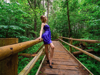 Young teenager girl dressed in stylish blue high fashion dress posing on a wooden bridge in a forest park. Prom time. Model wears design clothes. Glamour and elegance concept.