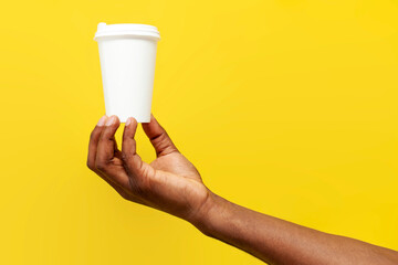 african american man's hand holds white paper cup of coffee on yellow isolated background