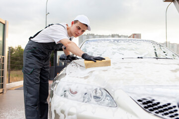 young guy in uniform car wash worker washes car with sponge with foam