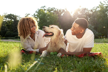 african american happy couple lie together with dog in park in summer, man and woman hug and love retriever