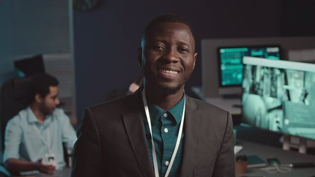Medium portrait of young African American cybersecurity specialist smiling at camera showing his badge standing in dark cyber security agency, working there with diverse colleagues