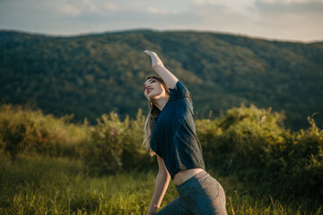 Fototapeta premium A young woman gracefully extends her arms in a picturesque yoga pose, surrounded by lush greenery under a clear blue sky.