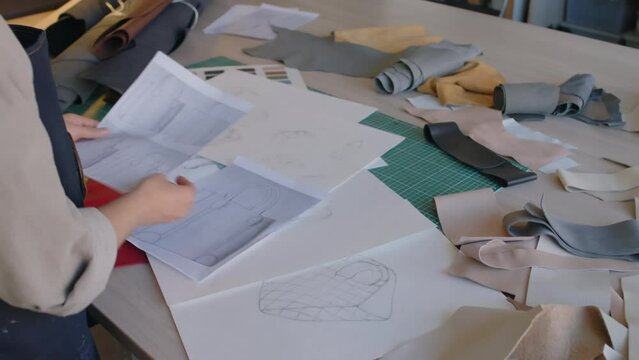 Close-up over-shoulder shot of unrecognizable female pattern cutter looking at fashion drawings and sketches and choosing leather pieces for handbags