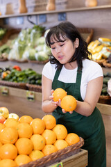 Young woman in apron working in salesroom of greengrocer, setting out fruits on shelves