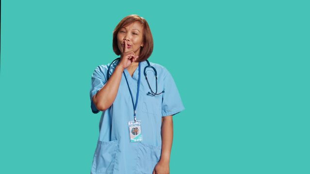 Portrait of stern serious asian nurse shushing at camera, bothered by excessive noise. Strict healthcare manager holding quiet gesture finger to lips, isolated over blue studio background