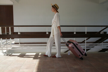 Tourist woman with sun hat carrying wheeled luggage walking in resort loking for her room. Travelling, vacation, leisure, summer holiday, solo traveling concept. - obrazy, fototapety, plakaty