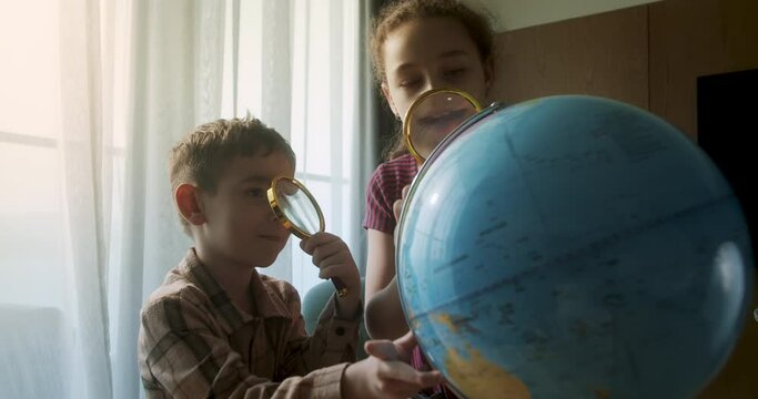 Kids in morning light sunset.Cute little Children with Globe and magnifying glass, technology for schoolboy, child looks at globe. Children is studying while sitting at home in rays sunset in room. 4K