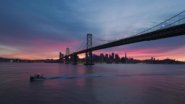 Aerial shot of small boat sailing under San Francisco Bay Bridge with scenic downtown on cinematic background with pink blue sunset sky above. Sark silhouettes of bridge and San Francisco downtown USA