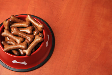 Red bowl with bone shaped dog cookies on wooden table, space for text