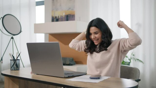 Happy latin woman saying YES looking at laptop. Excited smart hispanic businesswoman in casual sweater sitting at home office desk raising arms and dancing in celebration of successful job promotion